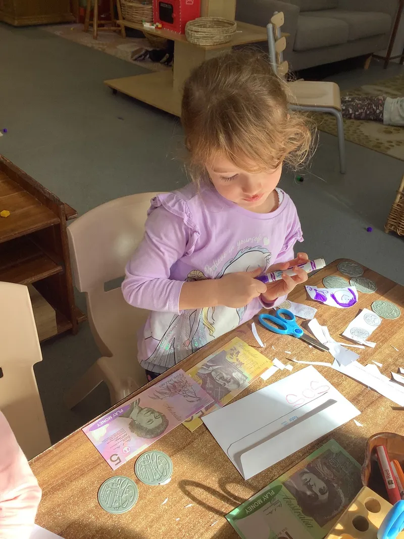 A preschool girl cutting out a paper coin at a table spread with banknotes, coins and worksheets.