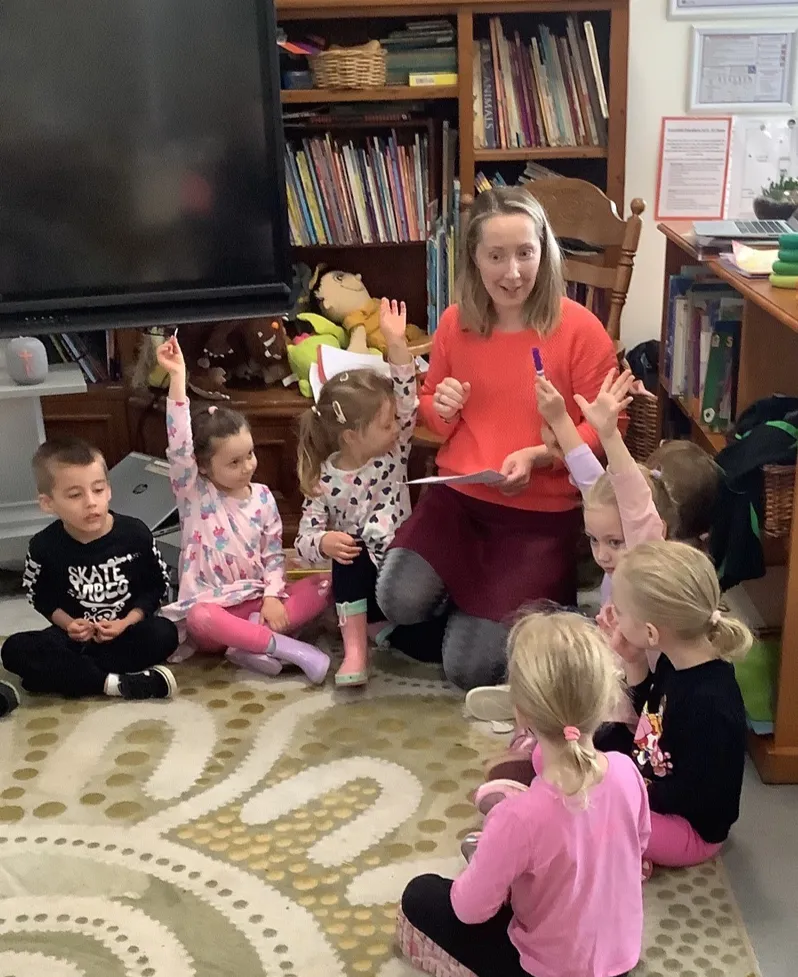 Preschoolers sitting on the floor with their hands up, answering the educator during a money workshop.