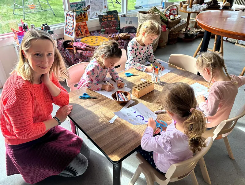 The educator seated with five preschoolers around a table, working through coin worksheets together.