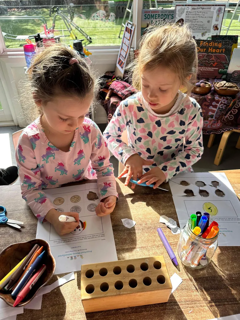Two preschoolers standing side by side, matching real coins to the pictures on their worksheets.