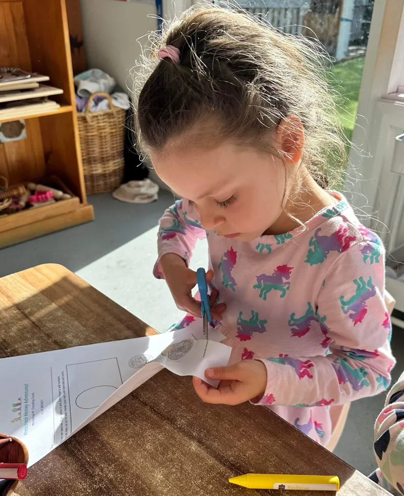 A preschooler concentrating as she cuts a paper coin out of her worksheet with scissors.