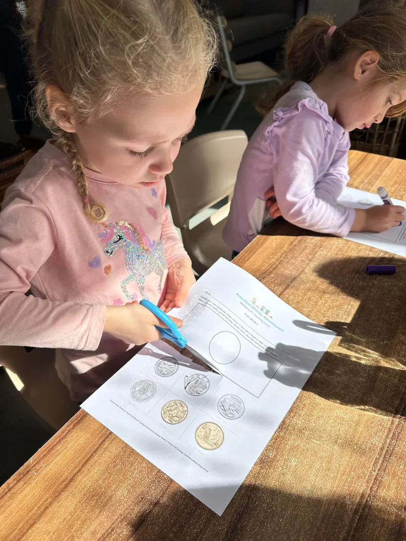 A preschool girl cutting out coins while another child colours her own money worksheet beside her.