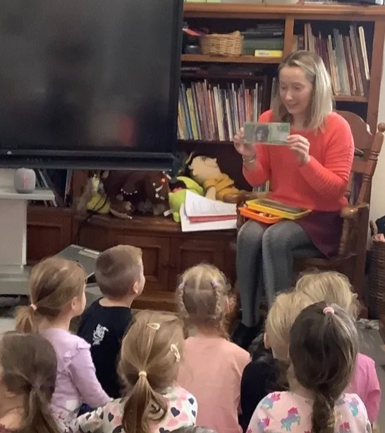 The educator holding up a banknote for a group of preschoolers gathered on the floor in front of her.