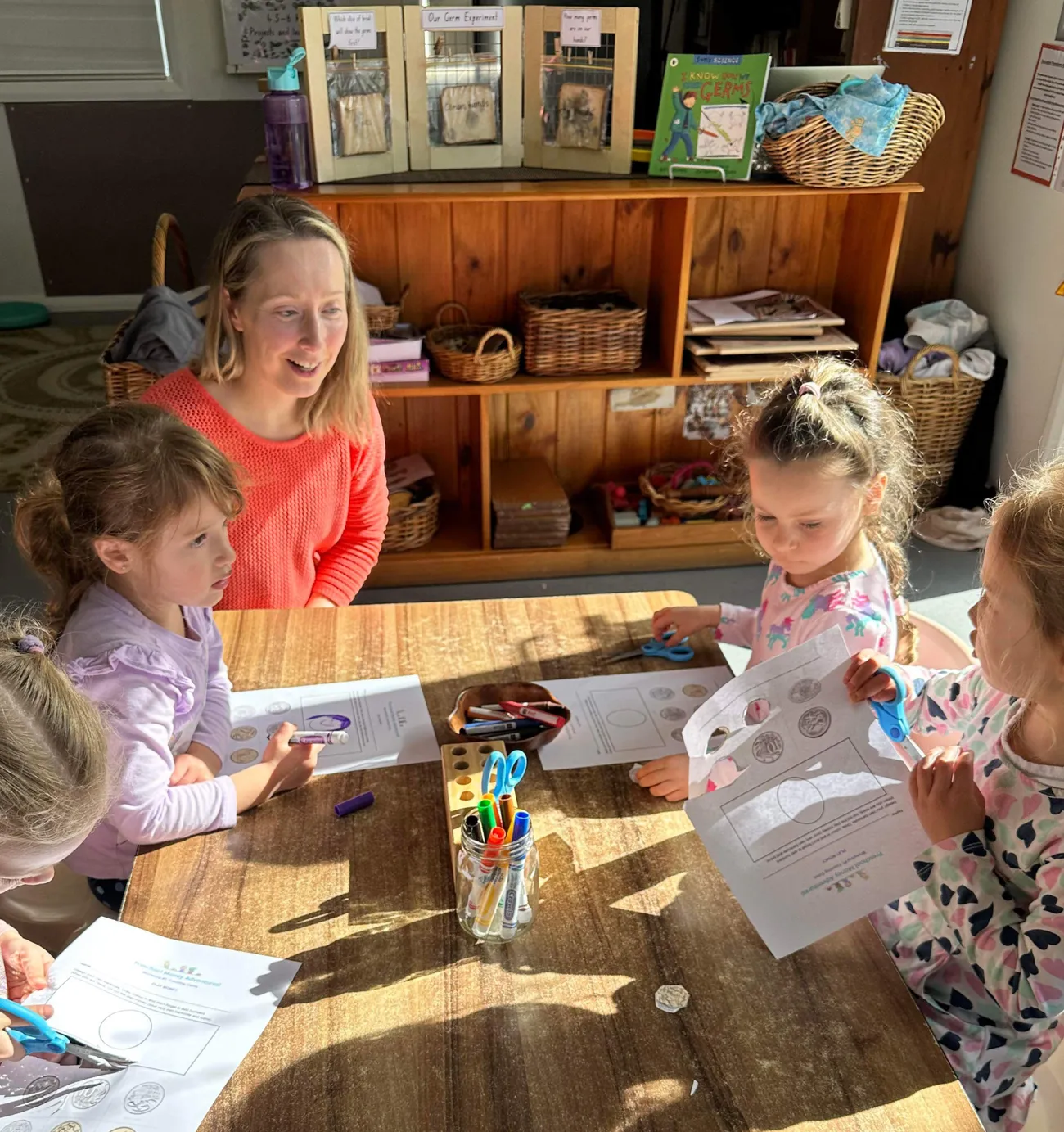 Four preschoolers around a wooden table with the workshop educator, sorting coins and working on money worksheets.